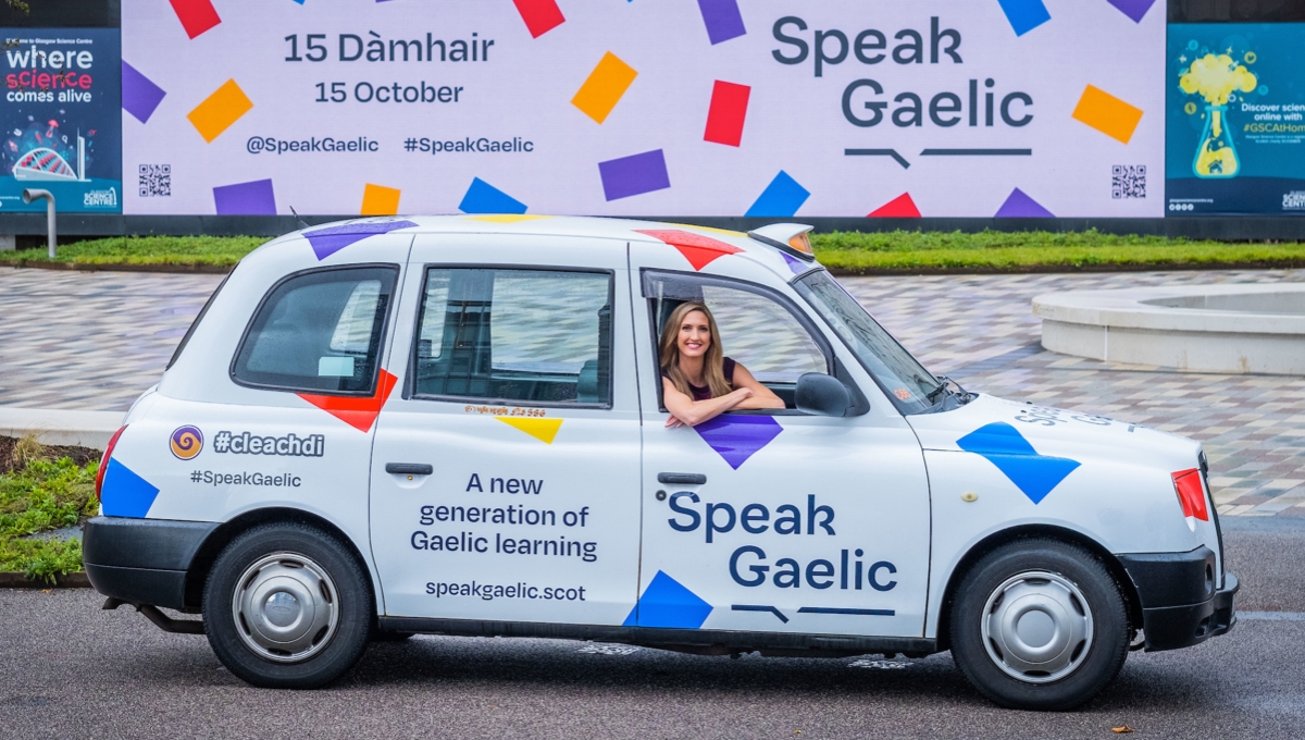 Joy Dunlop sits in the driver’s seat of a white SpeakGaelic branded taxi in in front of a SpeakGaelic billboard