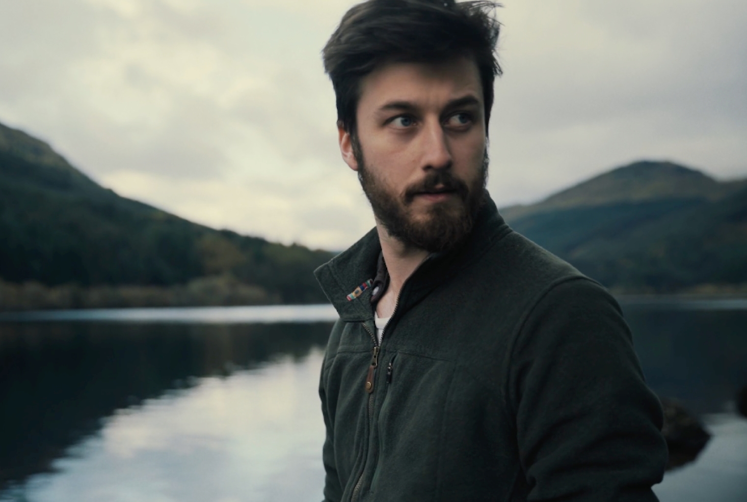 A photo of a man looking to the right of the camera with a loch behind him.