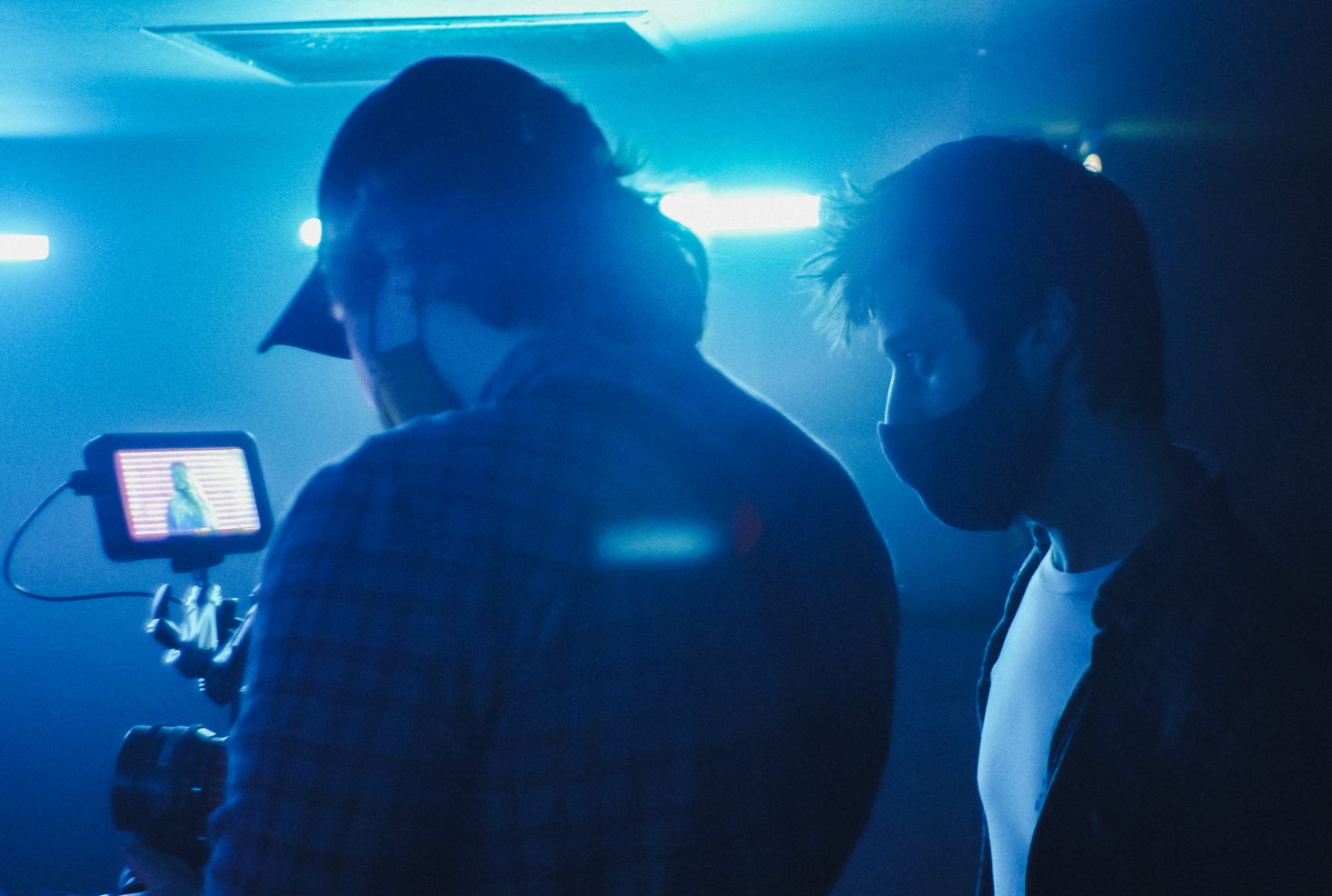Two midshot masked figures look into a mobile screen on a tripod.