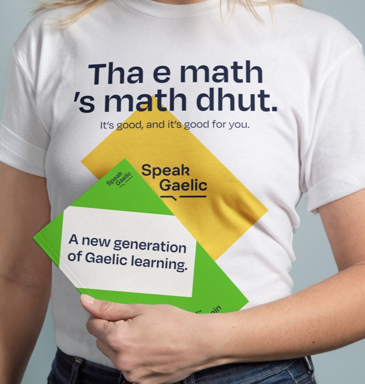 Someone wearing a SpeakGaelic t-shirt featuring a Gaelic proverb holding a SpeakGaelic book
