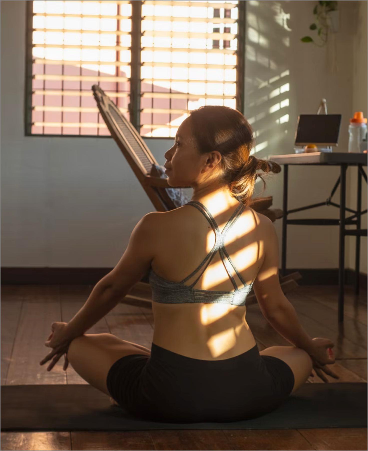 A photo of a woman in Easy Pose (Sukhasana) on a yoga mat. She sits facing away from the camera and her face is turned to the left. Light streams, through venetian blinds, onto her back