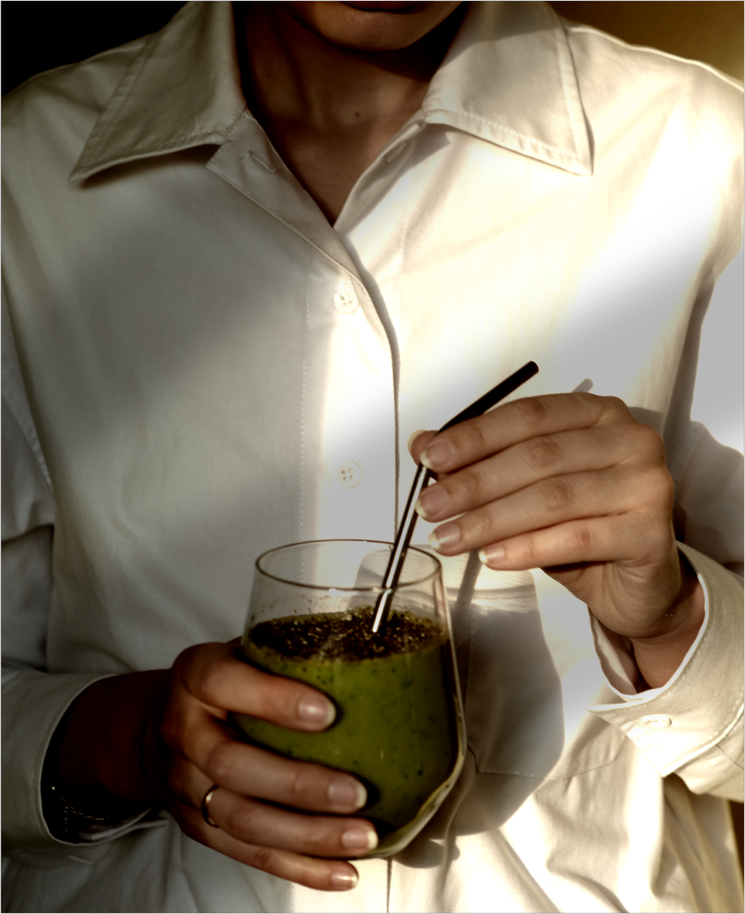 A picture of a torso in a white shirt with the person’s right hand holding a glass of green shake with a metal straw in it. Their left hand rests at the bend of the straw.