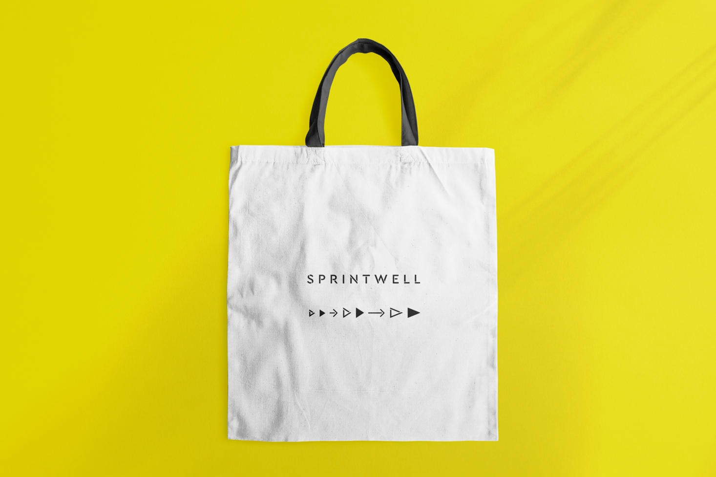 A white Sprintwell-branded tote bag with black handles with text “Sprintwell” and iconography sits on a yellow background.