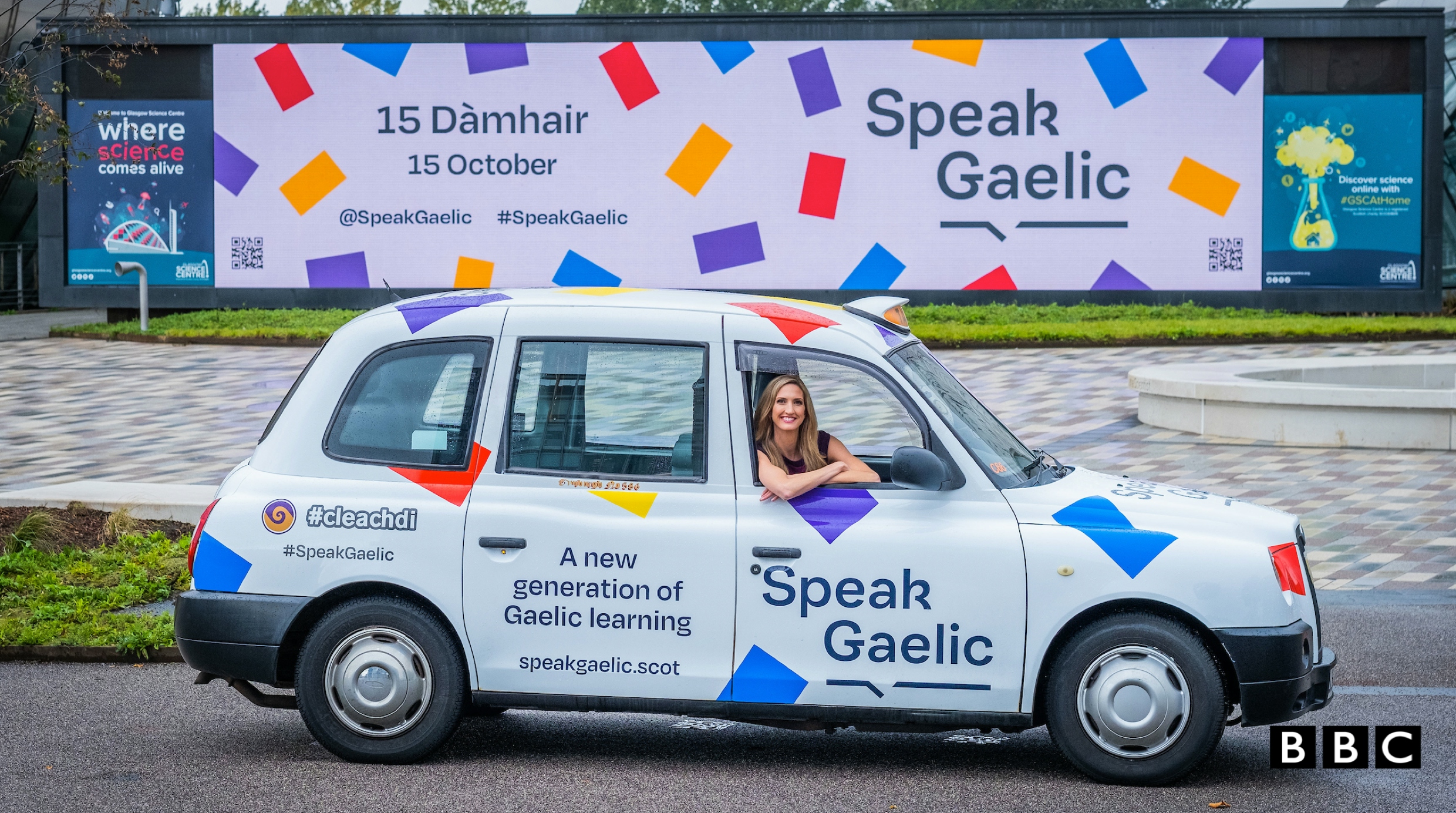 Joy Dunlop sits in the driver’s seat of a white SpeakGaelic branded taxi in in front of a SpeakGaelic billboard