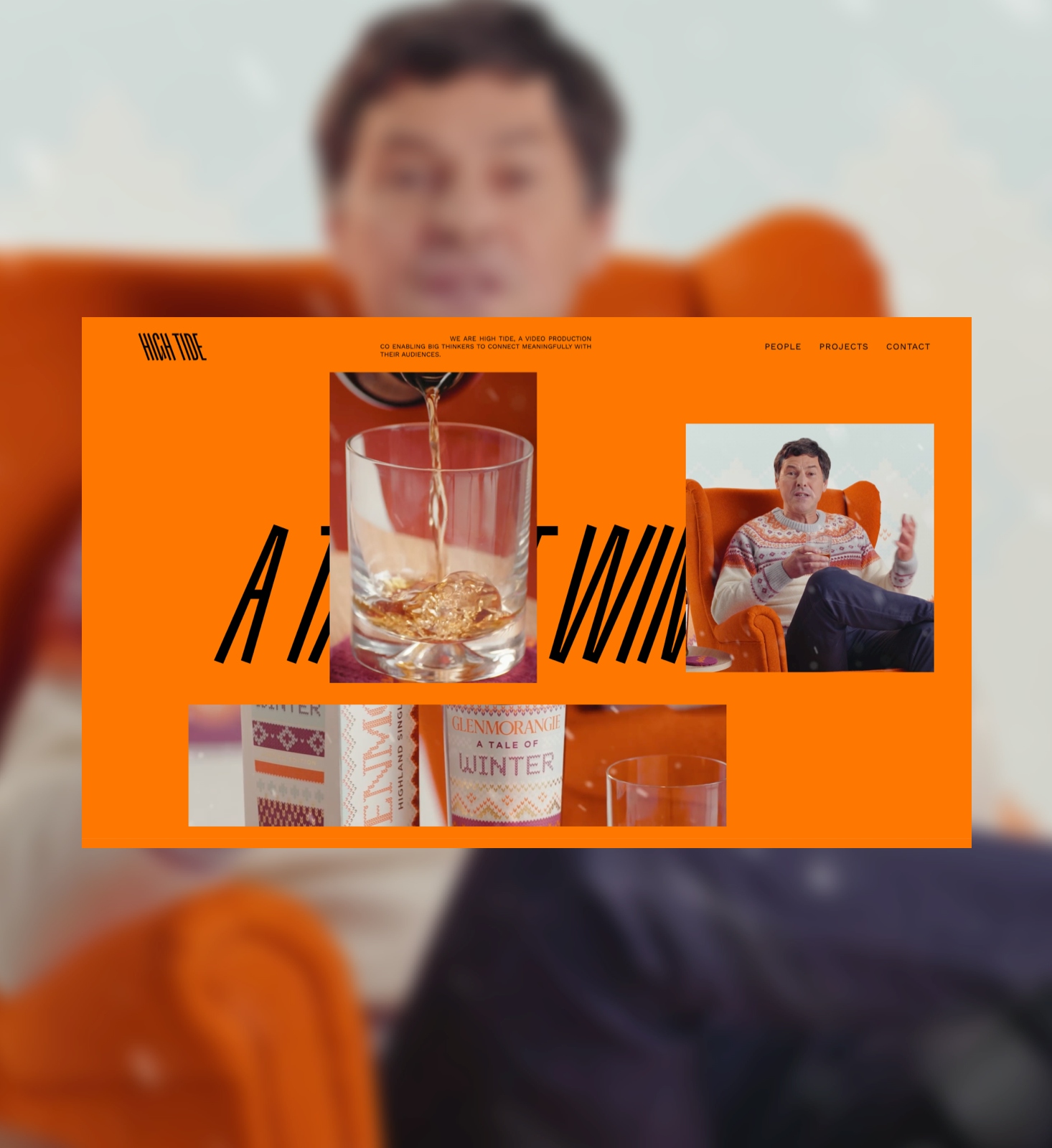 A blurry image of a man sitting on an orange armchair has an orange landscape webpage on it. The webpage has black letters on it and in the left half has a glass of whisky, to the right there is an image of a man sitting on an orange armchair. To the bottom left of the page is a panoramic image of a box of whisky, a bottle and a glass.