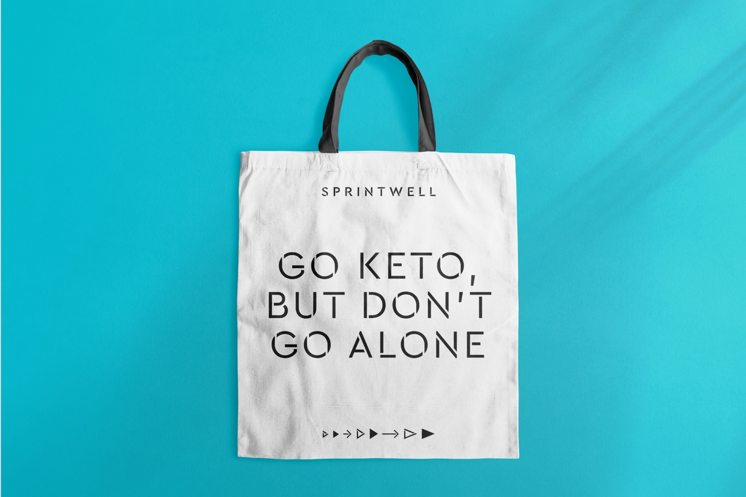 A white Sprintwell-branded tote bag with black handles with text “Go Keto but Don’t go alone” and iconography sits on a teal background.