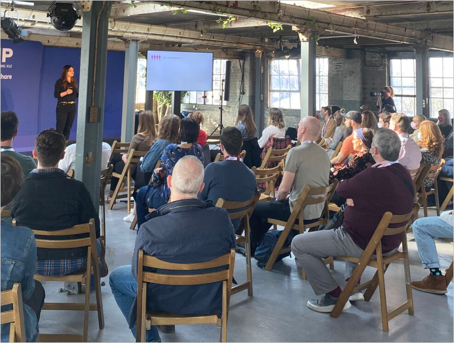 A photo of a talk being given; a crowd of approximately 40 attendees on wooden folding chairs facing a stage with a speaker and large monitor. Event is taking place in a refurbished industrial space, with exposed brick walls and steel support pillars.