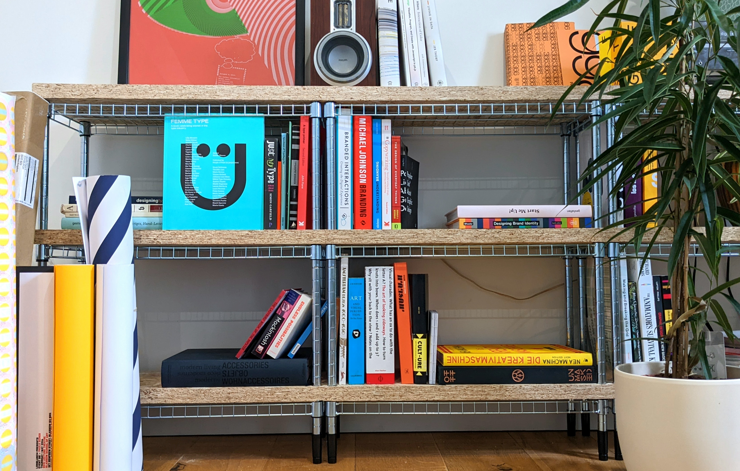 A set of metal shelves, with various colourful design books and a potted plant